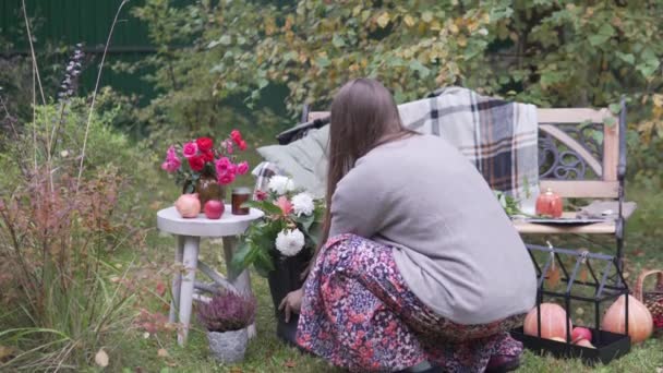 Kobieta zdobi ławeczkę i stół w ogrodzie z kwiatami i dyniami na wakacje. — Wideo stockowe