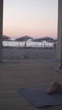 Gün batımında plaj yoga seansı, beyaz kadın ahşap pavyonun altında diz çöküp diz çökerek dans ediyor.