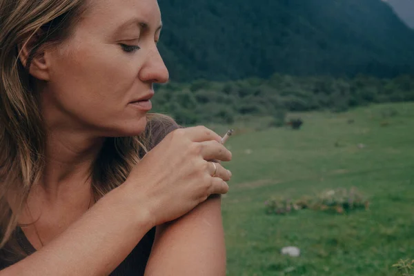 Yürüyüşten sonra omzunu kontrol eden bir kadının yakın çekimi, dağ çayırı arka planı, düşünceli ifade, sıcak doğal ifade