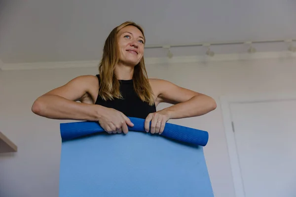 Elinde mavi minder tutan gülümseyen bir kadın. Evde yoga seansı hazırlıyor. Spor koçu rolü, yumuşak doğal ışık.,