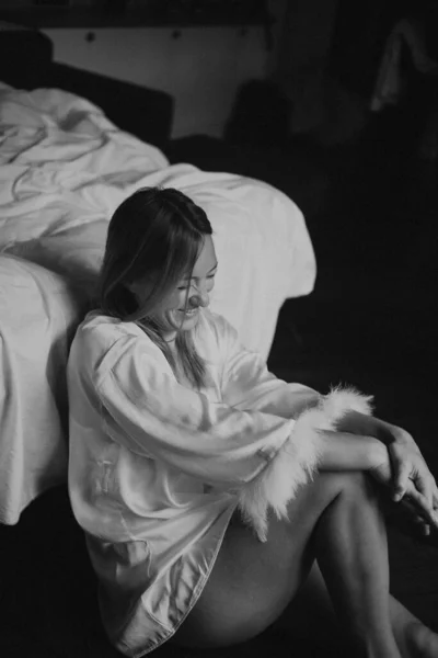 Kadın yatak odasında yerde oturuyor, omuzlarının üzerine yumuşak bir bornoz örtülmüş, güneş ışığı çıplak ayaklarda, düşünceli.
