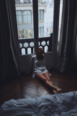 Beyaz kadın uzun pencereden şehre bakıyor. Bacakları açık, sırtı perdeye dönük.,