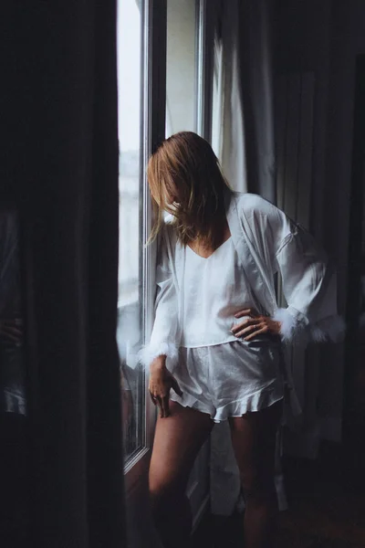 Pencere kenarında duran saten bornozlu beyaz kadın, elleri kalçasında, tüylü manşet ayrıntıları, yumuşak sabah ışığı,