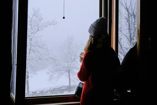 Örgü şapkalı ve eşarplı sıradan bir kız sıcak bir içecekle elinde kupayla pencereden kışın karlı manzaraya bakıyor. Rahat bir ev. Dağda Tatil