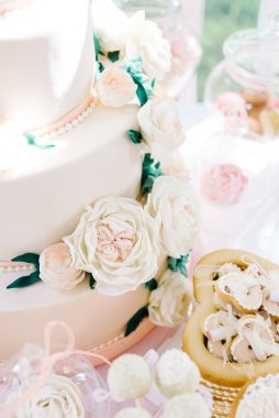 Çikolata. Çiçeklerle süslenmiş beyaz düğün pastası, çöl ve çöreklerle dolu bir şenlik masasının önünde duruyor.