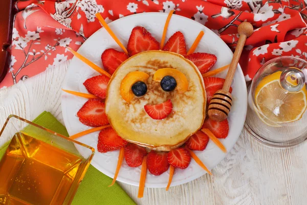 Childrens breakfast pancakes smiling face of the sun strawberry blueberry and apricot, cute food, honey — Stock Image Childrens breakfast pancakes smiling face of the sun strawberry blueberry and apricot, cute food, honey Stock Photo