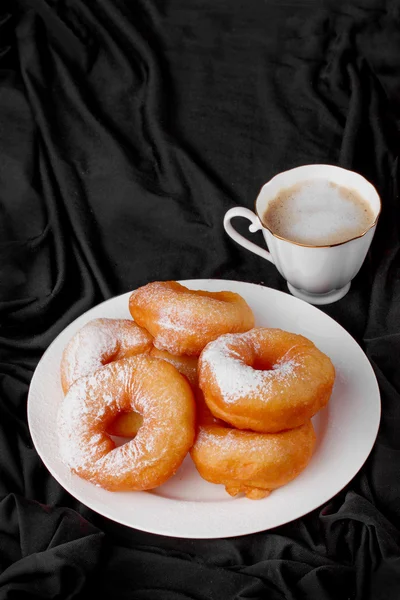 Donuts with powdered sugar and coffee on a black background drapery fabric — Stock Photo, Image