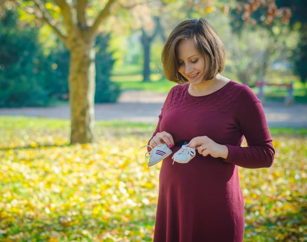 Frumoasă femeie însărcinată care ține pantofi pentru copii în parcul de toamnă — Fotografie, imagine de stoc