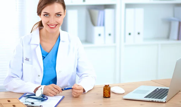 Hermosa joven sonriente doctora sentada en el escritorio y escribiendo. — Foto de Stock