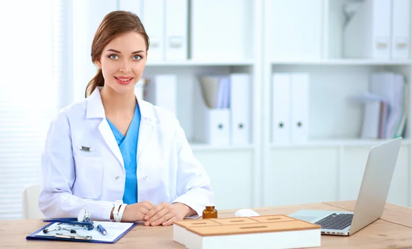 Schöne junge lächelnde Ärztin sitzt am Schreibtisch und schreibt. — Stockfoto