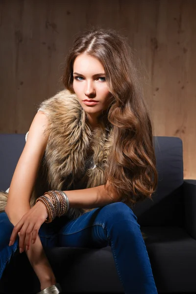 Portrait of elegant woman sitting on black sofa wearing a blue jeans and fur vest — Stock Photo, Image