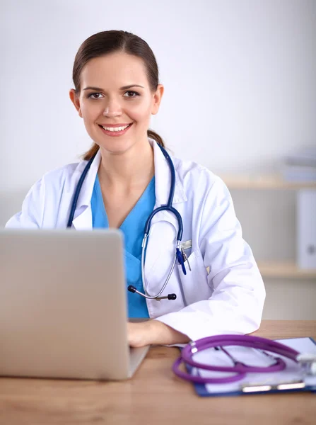 Mooie jonge glimlachende vrouwelijke arts zitten aan het bureau en schrijven. — Stockfoto