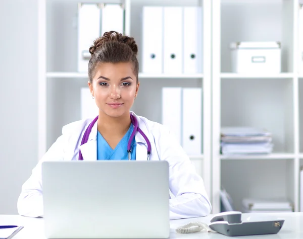 Mooie jonge glimlachende vrouwelijke arts zitten aan het bureau en schrijven. — Stockfoto