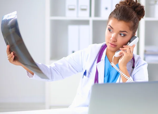 Médico femenino que mira la radiografía y habla por teléfono en el centro de diagnóstico, sentado en el escritorio — Foto de Stock