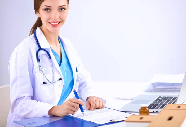 Hermosa joven sonriente doctora sentada en el escritorio y escribiendo. — Foto de Stock