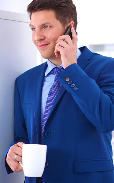 Hombre de negocios sonriente de pie y usando el teléfono móvil en la oficina —  Fotos de Stock