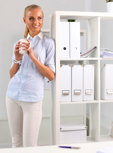 Junge Geschäftsfrau steht mit Tasse am Schreibtisch Stockbild