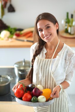 Mutfakta elinde sebzelerle gülümseyen genç bir kadın.