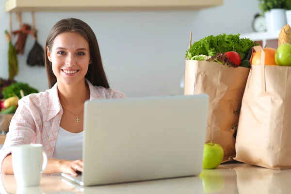 Smiling young woman with coffee cup and laptop in the kitchen at home — Stock Photo, Image