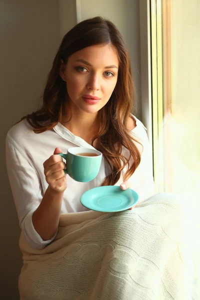 Jonge vrouw thuis zitten in de buurt van raam ontspannen in haar woonkamer lezen boek en het drinken van koffie of thee — Stockfoto