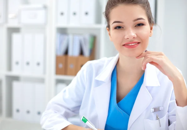 Mooie jonge glimlachende vrouwelijke arts zitten aan het bureau en schrijven. — Stockfoto