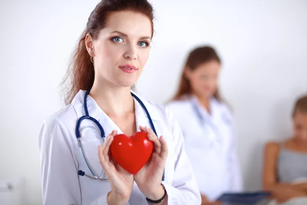 Mujer joven médico sosteniendo un corazón rojo, de pie sobre fondo gris —  Fotos de Stock