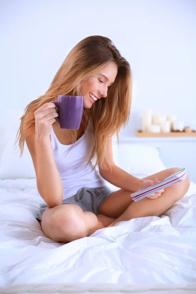 Mooie vrouw drinken een kopje koffie zittend op haar bed thuis — Stockfoto