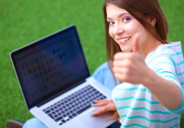 Laptoplu genç bir kadın yeşil çimlerde oturuyor.