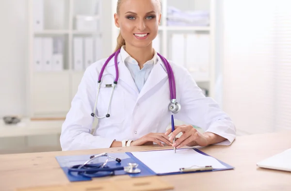 Hermosa joven sonriente doctora sentada en el escritorio y escribiendo. —  Fotos de Stock