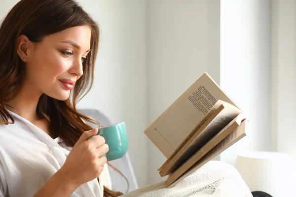 Fiatal nő otthon ül az ablak mellett pihentető a nappaliban olvasás könyv és kávézás vagy tea — Stock Fotó
