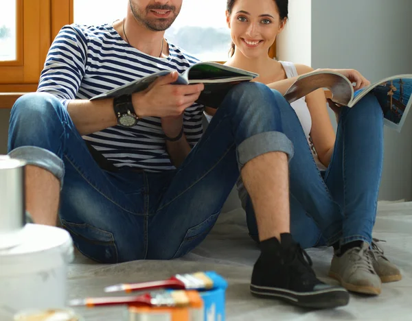 Casal atraente sentado no chão de casa olhando jurnal e sorrindo um para o outro . — Fotografia de Stock