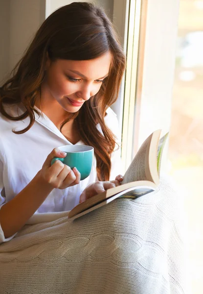 Tânăra de acasă stând lângă fereastră relaxându-se în sufrageria ei citind cartea și bând cafea sau ceai — Fotografie, imagine de stoc