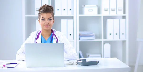 Mooie jonge glimlachende vrouwelijke arts zitten aan het bureau en schrijven. — Stockfoto