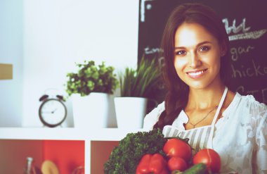 Mutfakta elinde sebzelerle gülümseyen genç bir kadın.