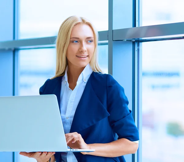 Donna d'affari in piedi contro la finestra dell'ufficio che tiene il computer portatile — Foto Stock