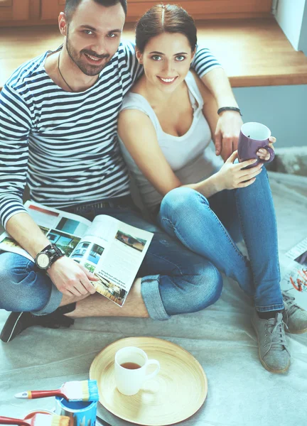 Casal atraente sentado no chão de casa olhando jurnal e sorrindo um para o outro . — Fotografia de Stock