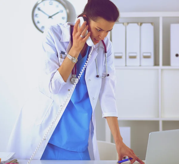 Young woman doctor in white coat at computer using phone — Stock Photo, Image