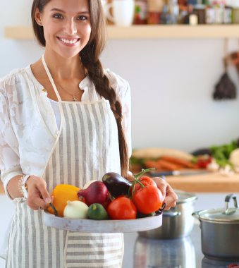 Mutfakta elinde sebzelerle gülümseyen genç bir kadın.