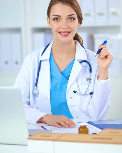 Hermosa joven sonriente médico femenino sentado en el escritorio y w —  Fotos de Stock