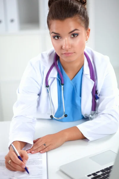 Hermosa joven sonriente doctora sentada en el escritorio y escribiendo. —  Fotos de Stock