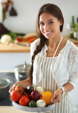Mutfakta elinde sebzelerle gülümseyen genç bir kadın.