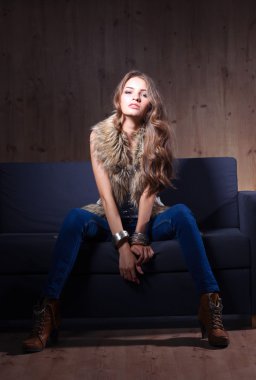 Mavi jeans giydiği siyah koltukta oturan zarif kadın portresi ve kürk yelek