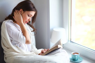 Evde oturan genç bir kadın oturma odasında oturmuş kitap okuyor ve çay ya da kahve içiyor.