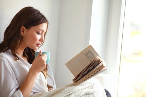 Mujer joven en casa sentada cerca de la ventana relajándose en su sala de estar leyendo libro y tomando café o té — Foto de Stock