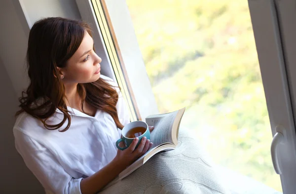 Ung kvinna hemma sitter nära fönstret koppla av i sitt vardagsrum läsa bok och dricka kaffe eller te — Stockfoto