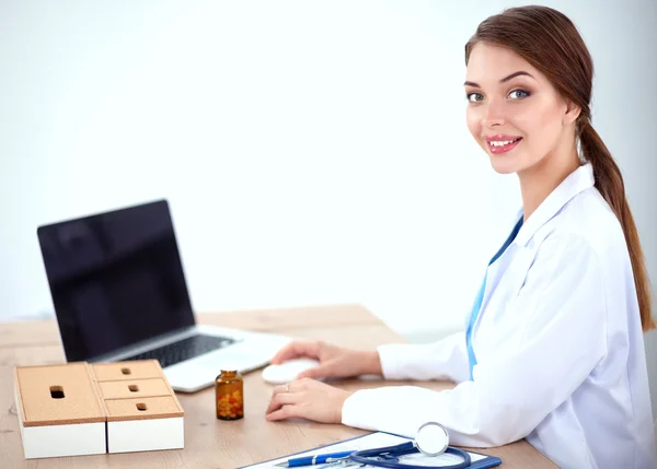 Hermosa joven sonriente doctora sentada en el escritorio y escribiendo. — Foto de Stock