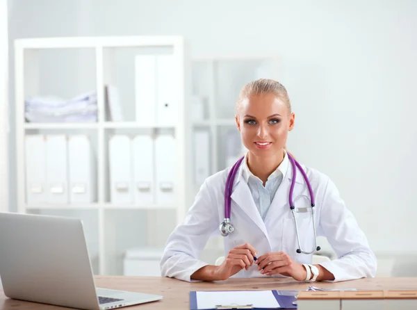 Mooie jonge glimlachende vrouwelijke arts zitten aan het bureau en schrijven. — Stockfoto