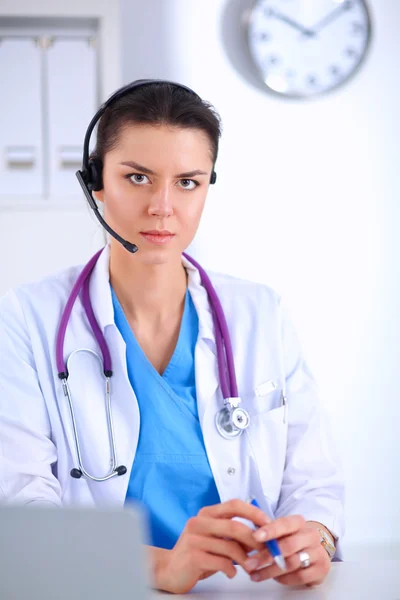 Lekarz w słuchawkach siedzi za biurkiem z laptopem na szarym tle — Zdjęcie stockowe