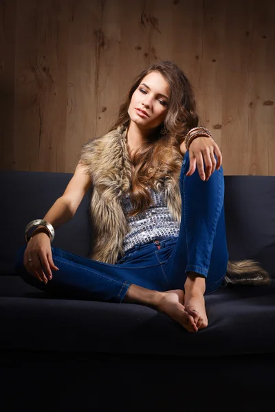 Ritratto di donna elegante seduta sul divano nero con jeans blu e gilet di pelliccia — Foto Stock