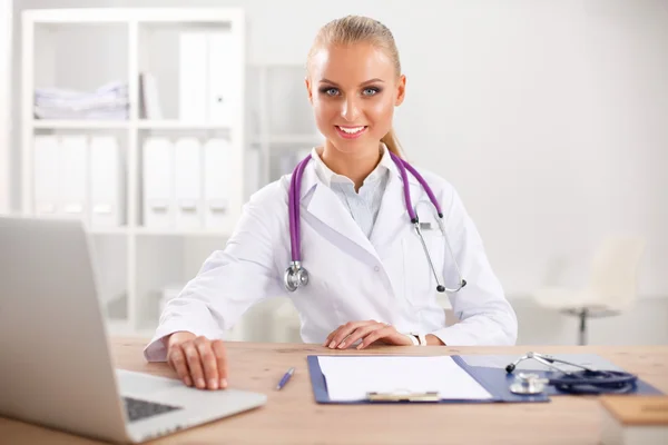 Mooie jonge glimlachende vrouwelijke arts zitten aan het bureau en schrijven. — Stockfoto